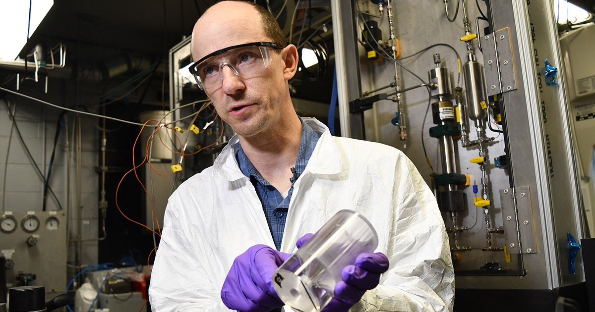 A professor wearing purple disposable gloves holds up a transparent combustion chamber for a small rocket engine