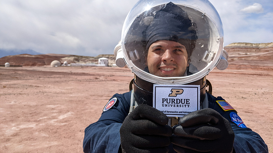 Julio Hernandez in a simulated space suit and holding up a Purdue patch, while participating in the Mars Analog Mission at Mars Desert Research Station