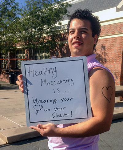 Julio Hernandez wearing a shirt with sleeves rolled up to show a heart drawn on his arm. He is holding a sign that reads, 