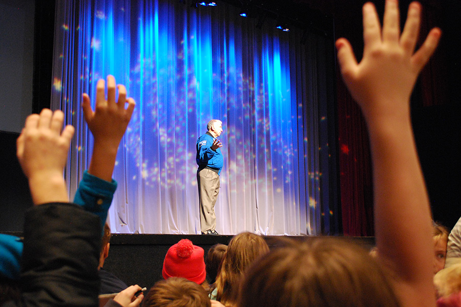 astronaut Charlie Walker standing on a stage. In the foreground, children's hands are raised to ask questions.