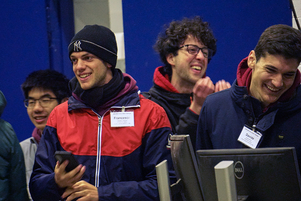 A group of people indoors, wearing coats and smiling. one person gesturing with his hands.