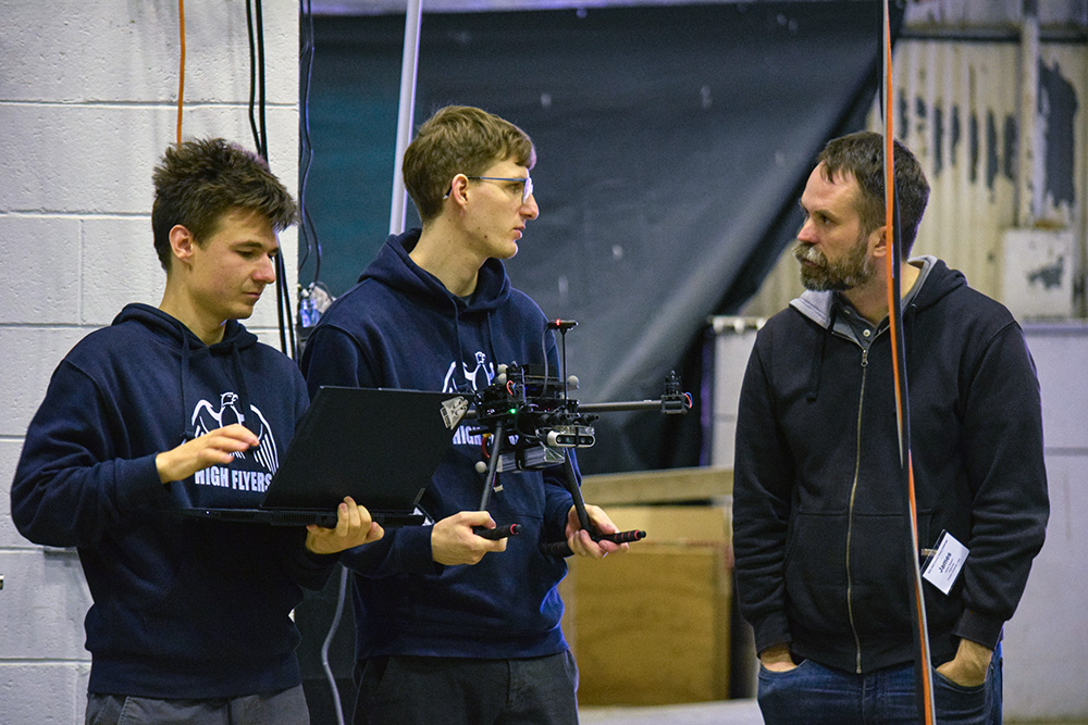 Two students, one holding a large quad-rotor drone, the other holding a laptop, speaking with a middle-aged man