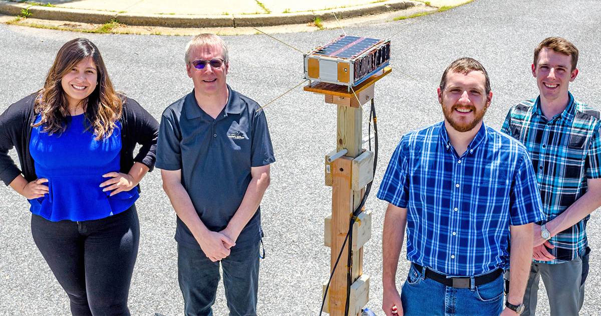 Group photo of students with Jim Garrison and the SNOOPI cubesat on a wooden stand