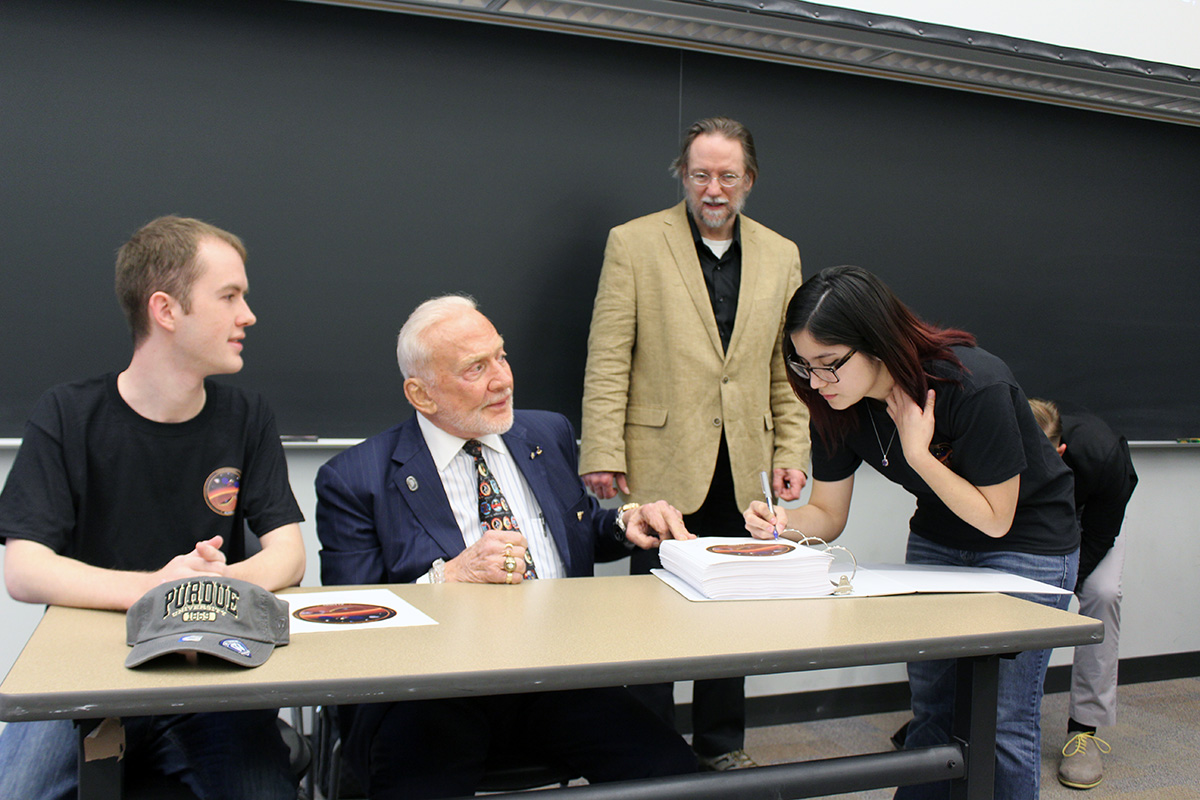 Longuski in the background as a student signs the first page of a binder on a table in front of astronaut Buzz Aldrin.