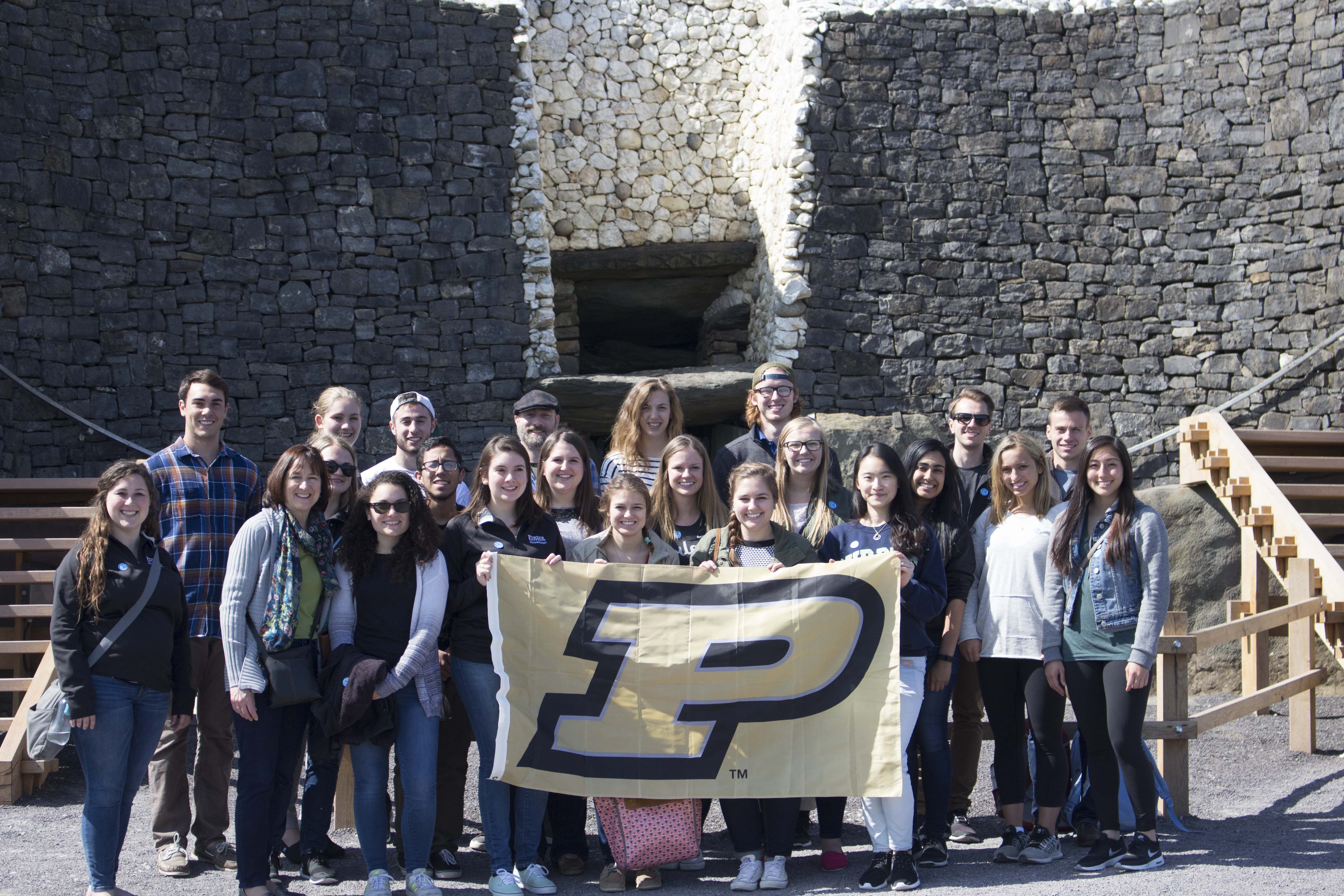 The students and advisors of the study abroad program holding a Purdue flag in front of Newgrange