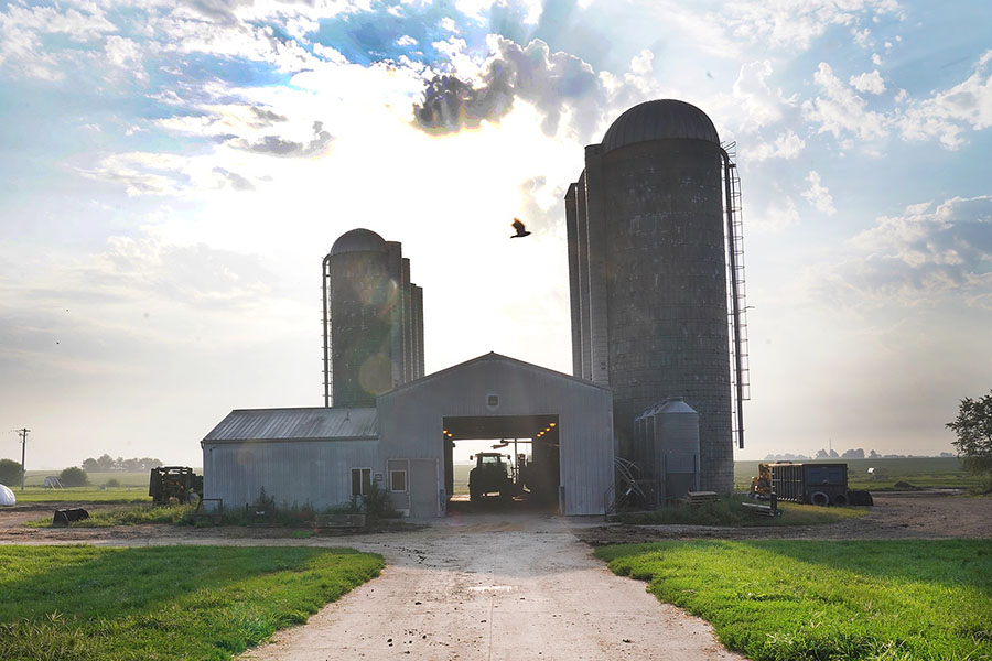 Photo of the Purdue Dairy Unit. Surrounded by green grass there is a barn with a tractor inside along with a few grain silos.