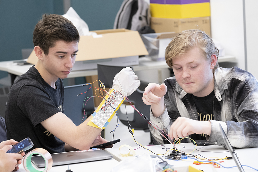 Two students are sitting at a desk working on a paper electronics project. The student on the left is wearing a glove connected to a variety of wires.