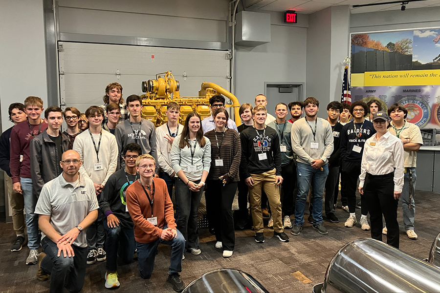 The READI program students and instructors pose for a group photo in front of a large engine at the Caterpillar plant.