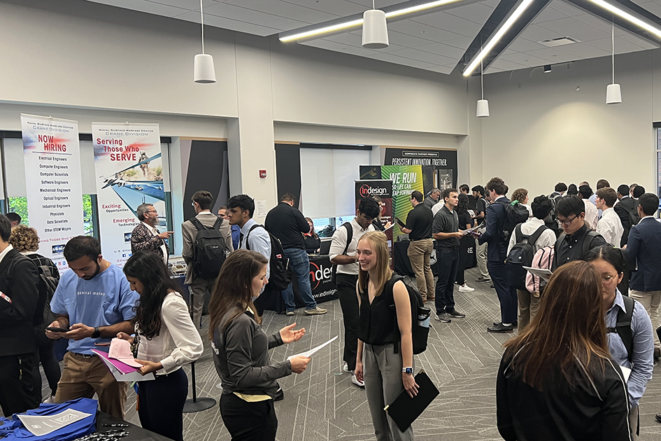 Students, dressed in business attire, are gathering in a large room to meet with companies who are recruiting. The company representatives are standing behind tables and banners to greet the students.