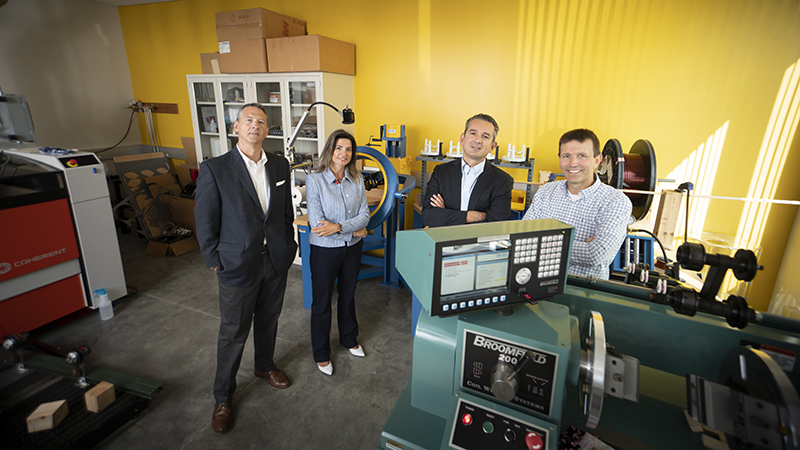 Purdue University engineers, from left, John Haddock, Nadia Gkritza, Dionysios Aliprantis and Steve Pekarek stand in the lab where they are testing technology they designed to enable electric vehicles to receive power from the road. There are several large instruments that surround them in the lab.
