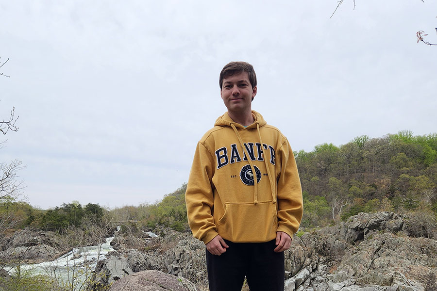 Student Ryan Gross poses for a picture while on a hike. He is wearing a yellow hoodie and is standing amongst rocks, trees, and a river.