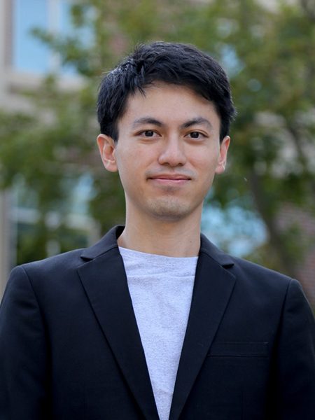 Kananart Kuwaranancharoen is posing for a portrait. He is wearing a t-shirt with a suit jacket. The background is blurred but contains a building and trees.