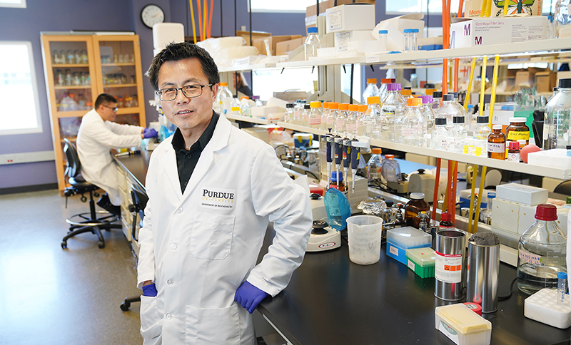 Professor W. Andy Tao stands in a lab. He is wearing glasses, a lab coat, and gloves. Behind him are various bottles and lab equipment.
