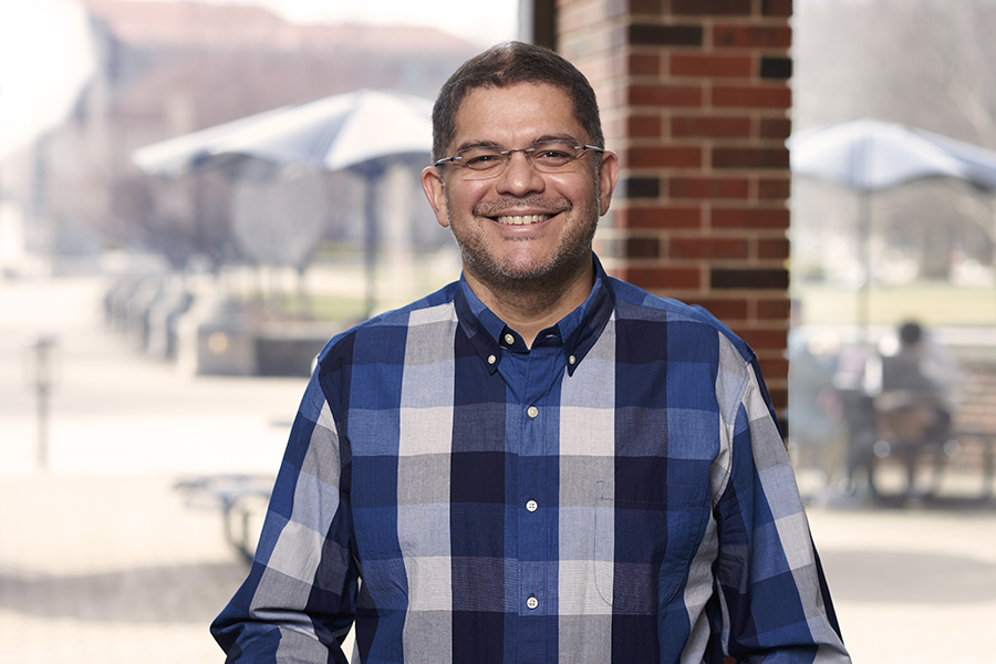 Professor Sunil Bhave stands in front of a window in the MSEE building for a portrait. He is wearing glasses and a blue and white plaid shirt.