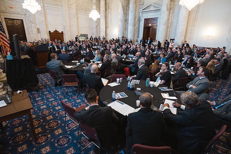 CHIPS for America summit attendees are seated at round tables in the right hand side of the image looking towards the left where panel speakers are sitting