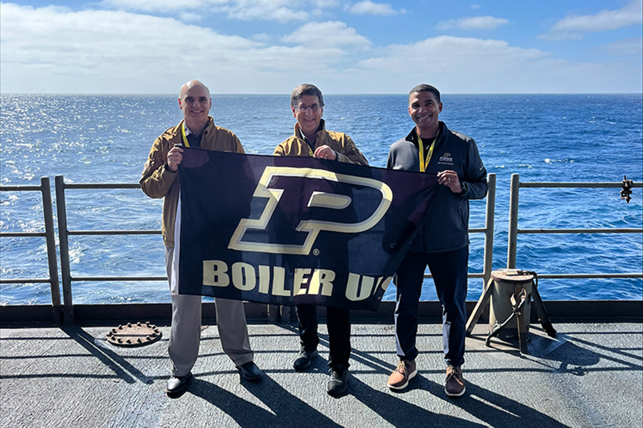 Dimitri Peroulis, Kelvin Gumbs, and Frank Dooley stand on the deck of the USS Nimitz holding a Purdue Boilermakers flag.