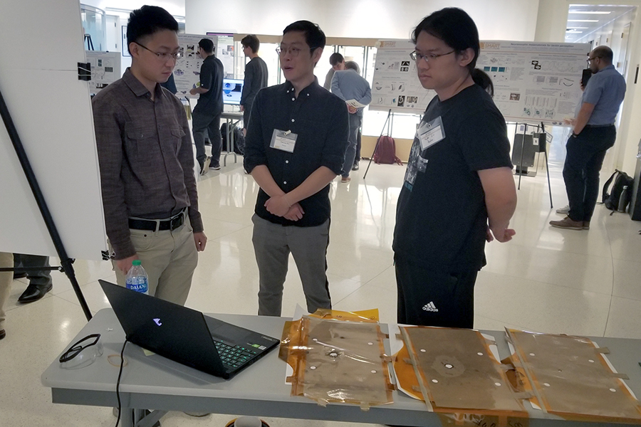 Yuzhang Zhu and Yufei Liu present their work to a third person standing between them. In the foreground, is a laptop and three of the impact sensors they designed with NASA.