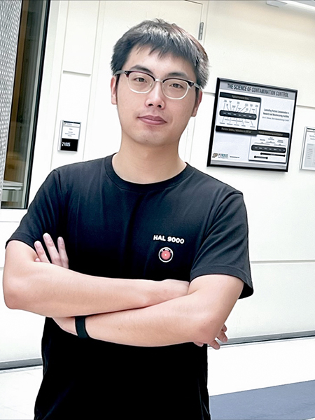 Xueji Wang is wearing a black t shirt and glasses. He is standing in front of a wall in Brick Nanotechnology Center.