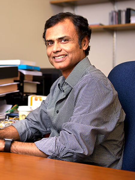 Kaushik Roy sits at his desk in his campus office. The desk is covered in various personal items and files.