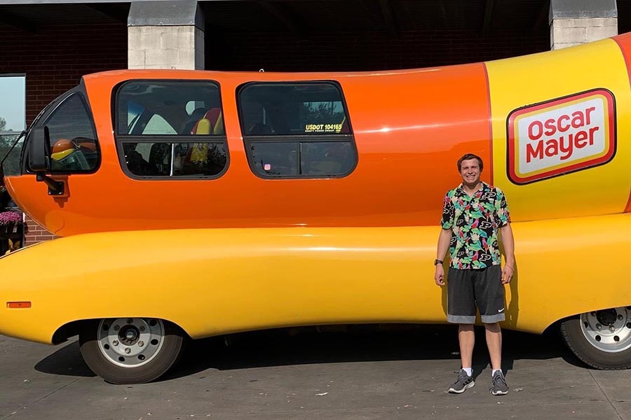 a male student, sam dlott, stands in front of the oscar mayer wienermobile which is a a long vehicle that resembles a hotdog