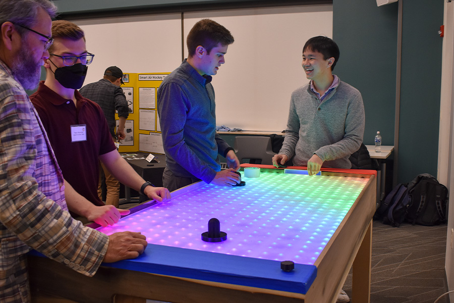3 students and professor look at the Smart Air Hockey Table which is lit up with many colored LED lights.