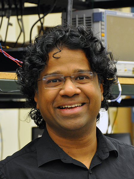 Portrait of Zubin Jacob in his lab. He is wearing a black polo while standing in front of some equipment.