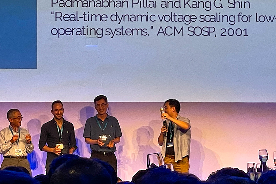 Purdue ECE Prof. Y. Charlie Hu accepts the 2023 ACM SIGMOBILE Test of Time Paper Award at ACM MobiCom 2023 on behalf of Abhinav Pathak, Ming Zhang, and himself. There is a gathering of people on a stage with a large projector screen behind them.