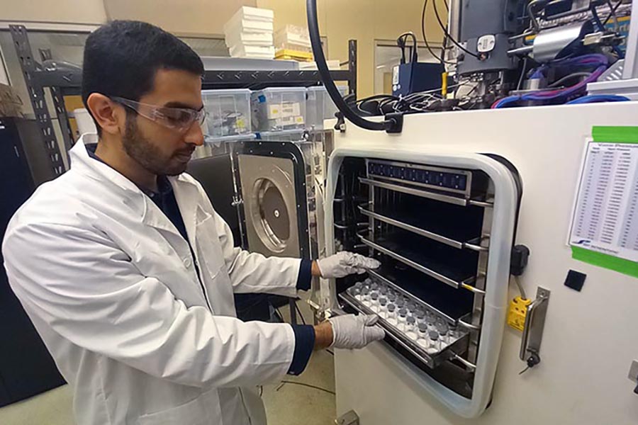Research Ahmad Darwish is placing a tray of vials into microwave lyophilizer which preserves vaccines. He is wearing a lab coat, gloves, and goggles.