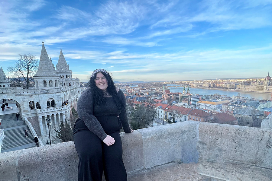 Amalia is sitting on a stone ledge. She is smiling and wearing all black and has a headband in her hair. In the background, there is a view of Fisherman's Bastion which is an old fortress in the city of Budapest. There is also a view of the Danube River.