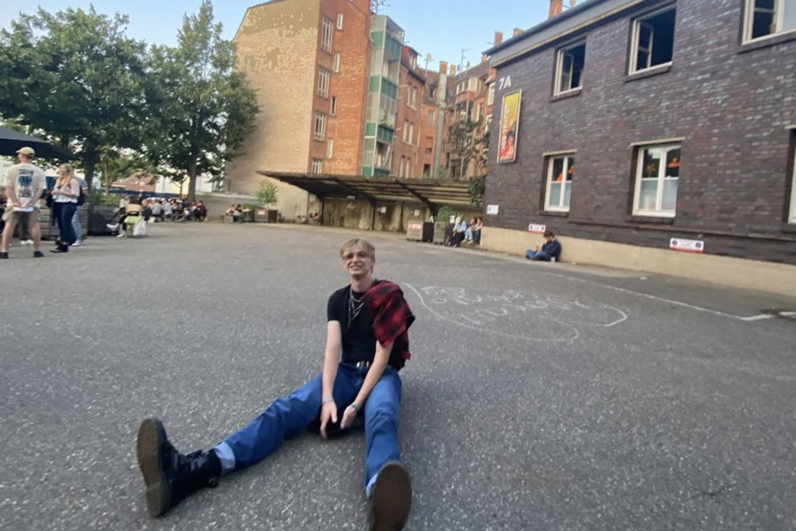 Brandon is wearing combat boots, jeans, and a black tshirt with a red planner thrown over his shoulder. He is stitting on the ground in a courtyard at a music festival in Stuttgart, Germany.