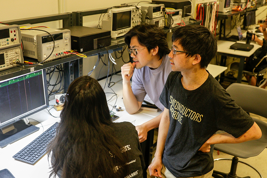 Three students are working at a desk in an electrical and computer engineering lab. They are looking intently at a monitor which is hooked up to lab equipment.
