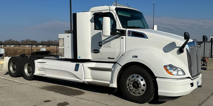 The semi truck equipped with the Cummins technology is parked on the side of a street with a fence.  The truck is painted white.