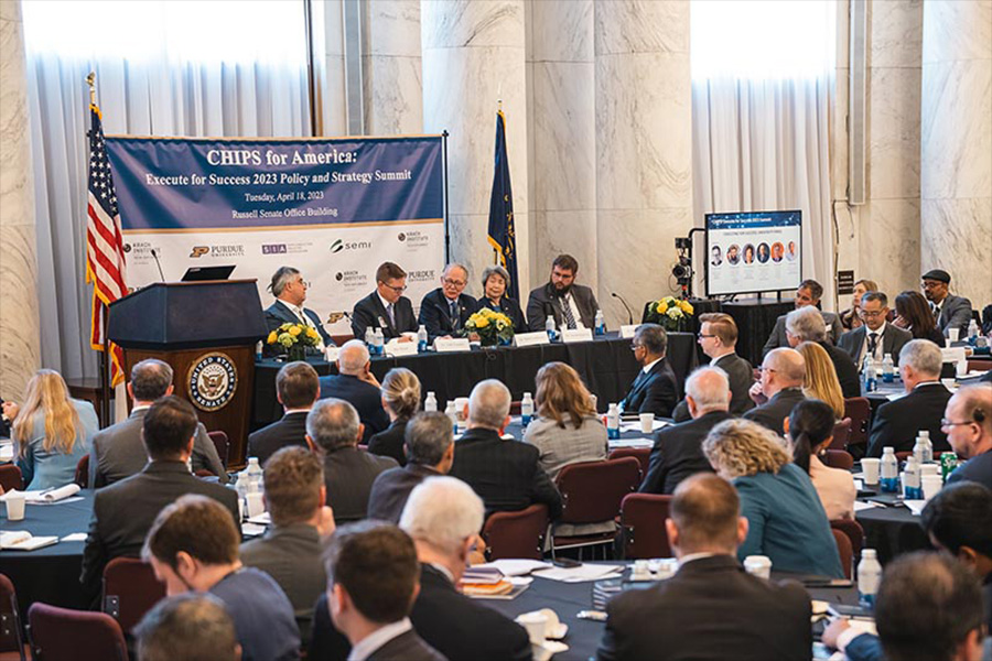 Purdue ECE Professor Mark Lundstrom sits on a panel discussion at a Chips for America summit. There is a banner and two flags behind the panel table, a podium to the left, and a TV screen to the right.