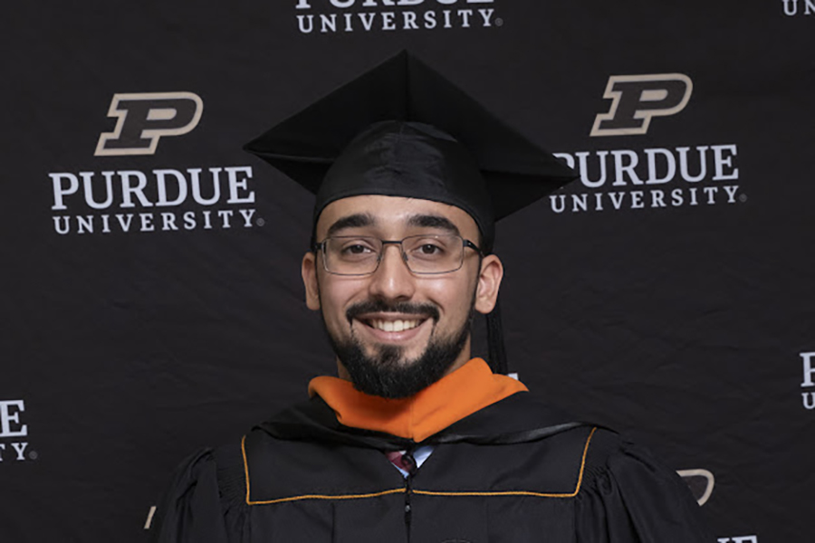 Omer, wearing his graduation regalia, poses for a portrait in front of a Purdue University backdrop.