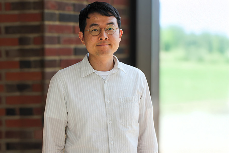 A bespectacled man poses in front of a rustic brick wall, highlighting his intellectual appearance and the wall's rough texture