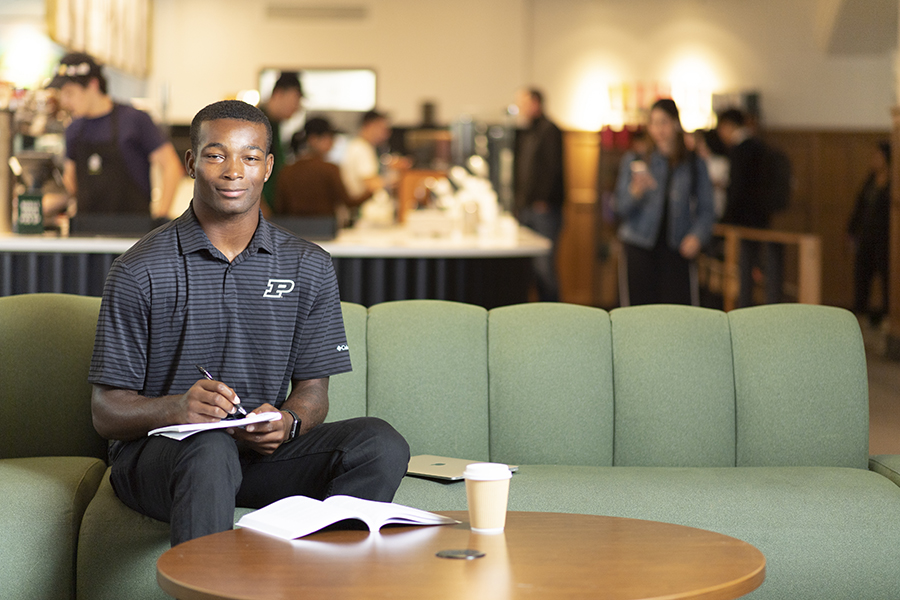 An African American student, wearing black pants and a Purdue polo, sit on a green couch with a tablet in hand. In the foreground a textbook and cup of coffee sit on a table.