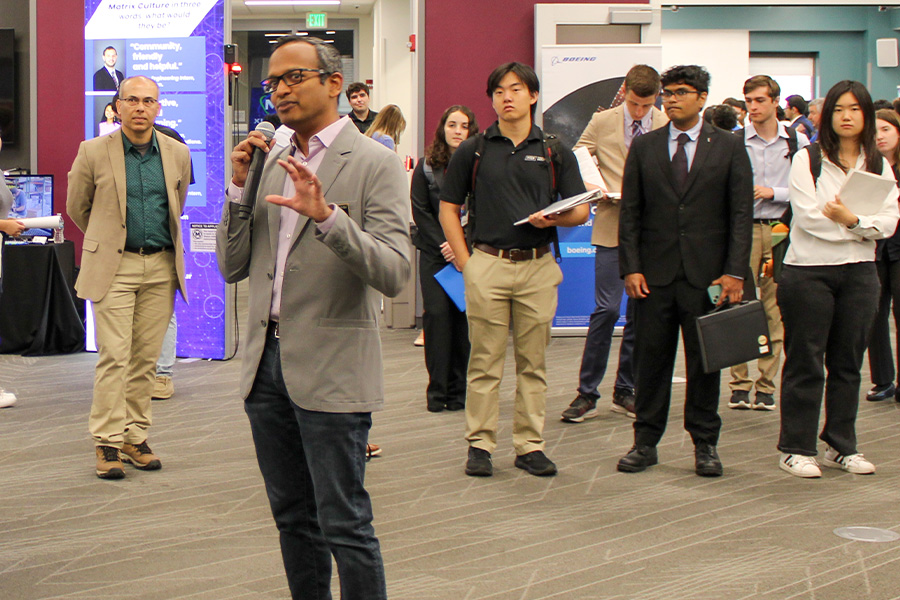 Man in gray blazer speaks into a microphone at a business event. Audience of diverse individuals in formal attire listens intently. Professional tone.