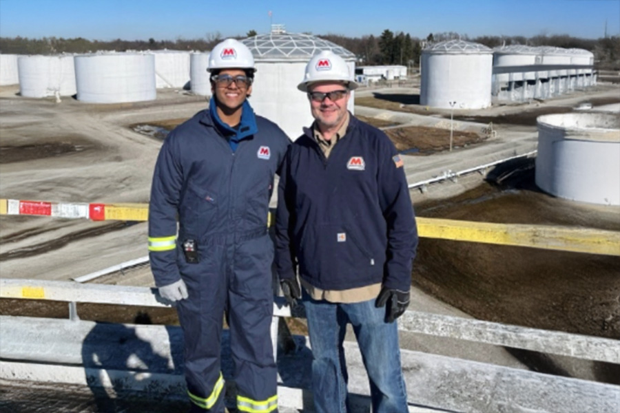 Two workers wearing safety helmets and gear smile in front of large industrial storage tanks under a clear blue sky, conveying professionalism and safety.
