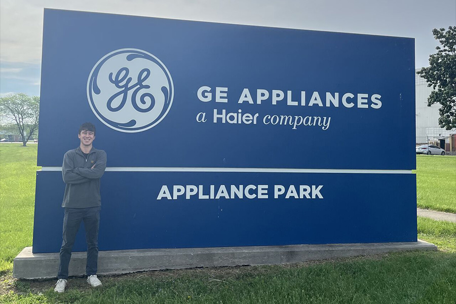 A person stands smiling with arms crossed next to a large blue sign for "GE Appliances, a Haier company, Appliance Park." The sky is overcast, creating a calm atmosphere.