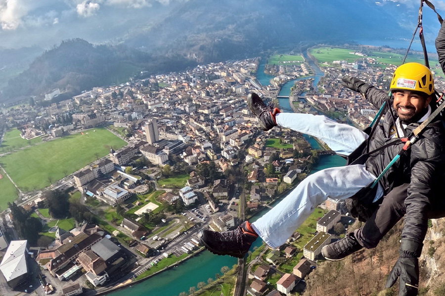 A person smiling widely while paragliding over a scenic town, wearing a yellow helmet and harness. Below is a sprawling landscape with buildings, green fields, and winding rivers, set against distant mountains. The scene conveys excitement and adventure.