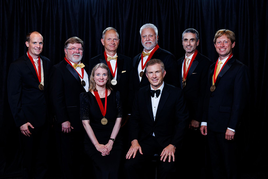 Eight people in formal attire pose against a black backdrop. Seven stand with gold medals on red ribbons, one person sits in front. The mood is celebratory.