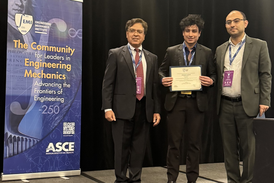 Three men pose at an engineering event; the center man holds a certificate, displaying pride. A banner reads, "The Community for Leaders in Engineering Mechanics."