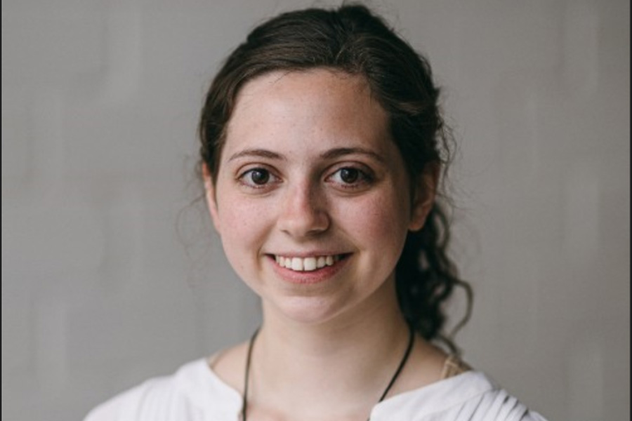 Smiling woman with long curly hair stands against a soft-focus neutral background, wearing a white blouse. Her expression conveys warmth and friendliness.