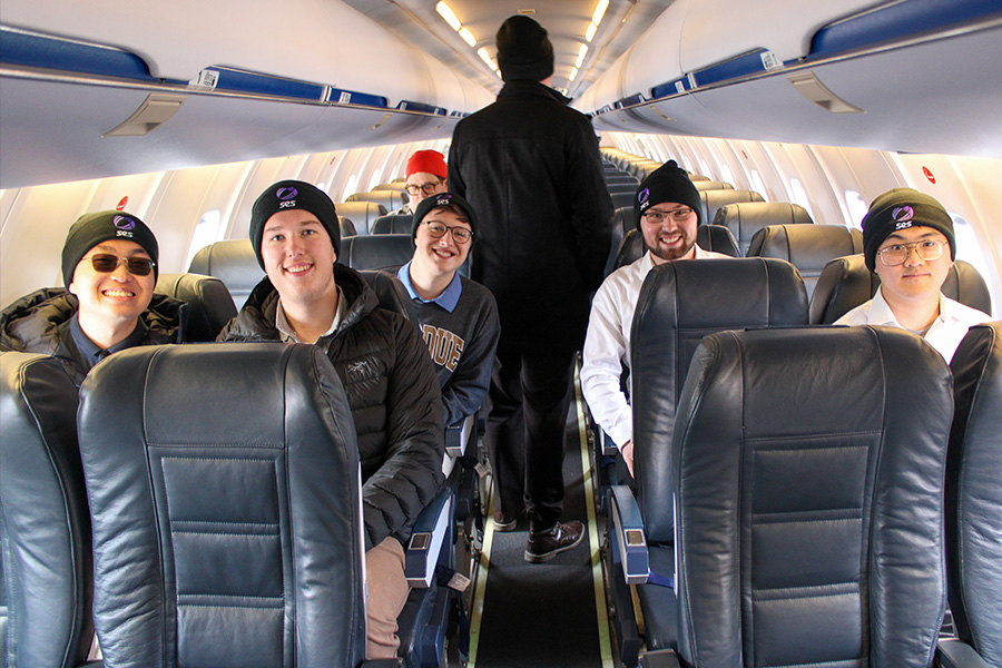 Six men stand smiling in front of a plane labeled SES on an airport tarmac. They wear casual and winter attire, creating a cheerful and relaxed atmosphere.