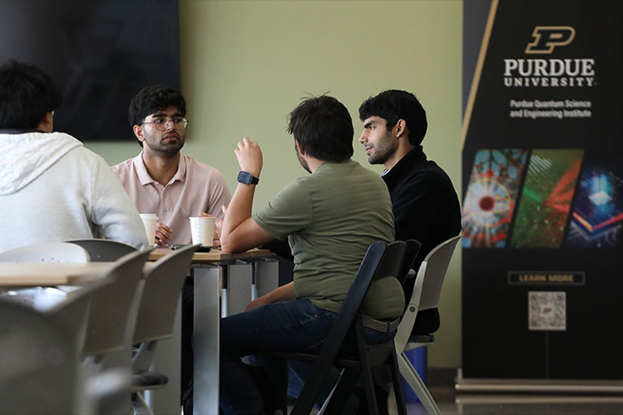 Four people in a discussion around a table, one wearing a green shirt gesturing. A Purdue University banner is visible, suggesting an academic setting.