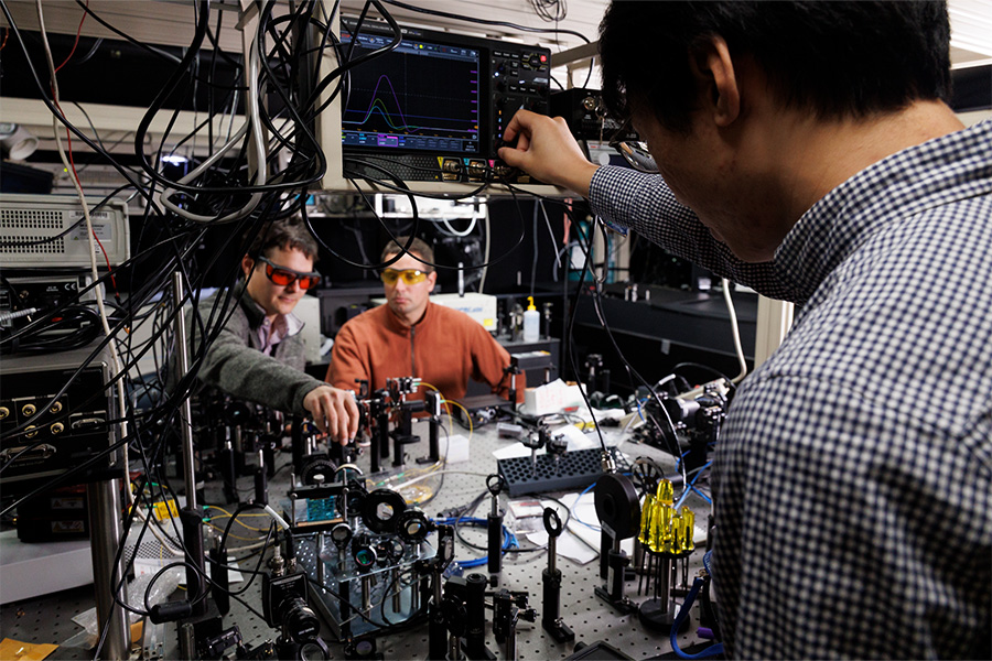 Three individuals work in a lab, surrounded by electronic equipment and cables. Two wear protective eyewear, while one adjusts a screen displaying graphs. The scene is focused and technical.