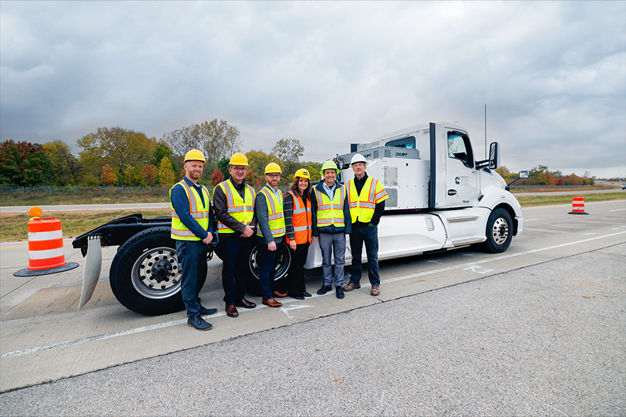 A group of researchers pose in front of a white semitruck while wearing bright yellow vests and hard hats.