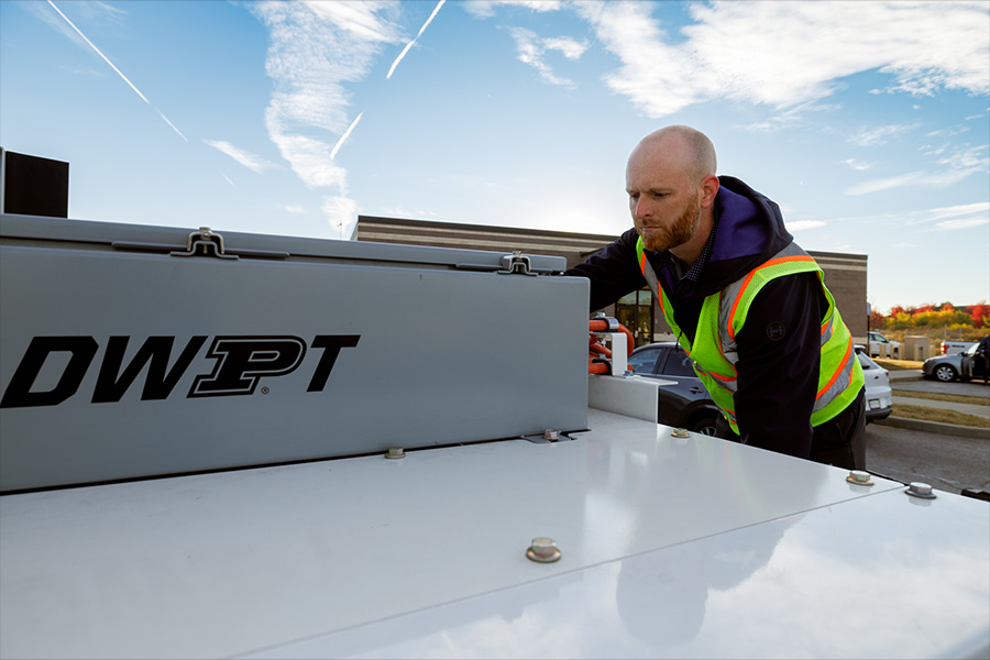 A man in a high-visibility vests leans over a trunk on the back of a semitruck. On the trunk is the text "DWPT."