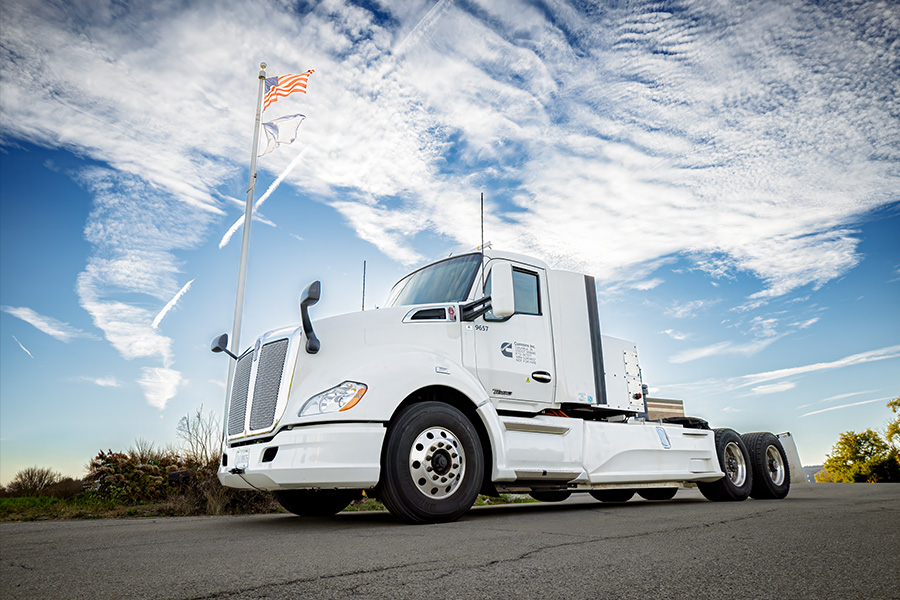 A white semitruck drives along a four-lane divided highway while two people on the shoulder of the road, wearing high-visibility vests and hardhats, take photos with their phones.