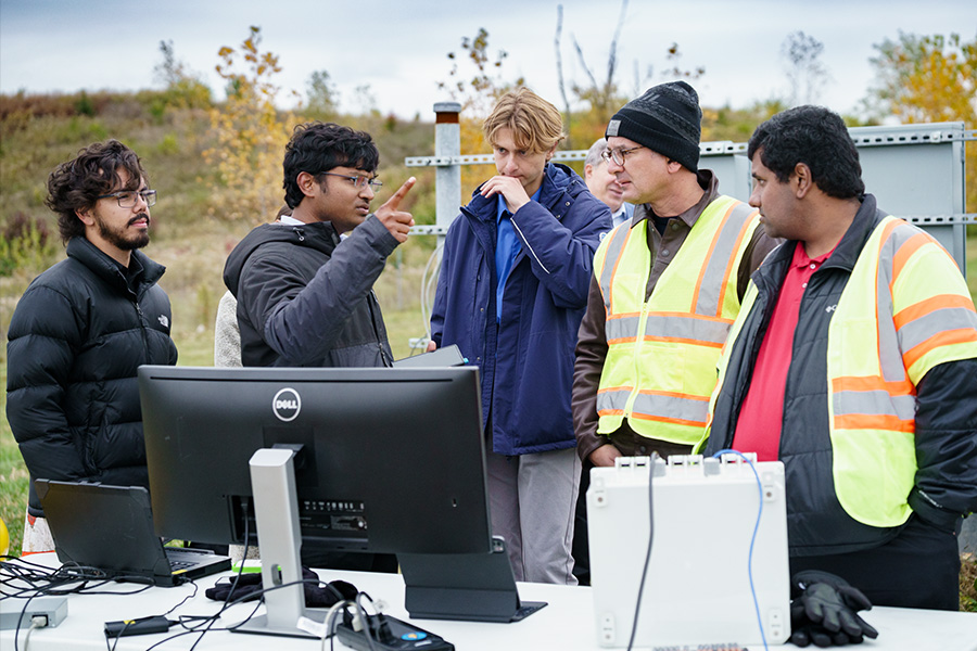 A group of students and researchers engage in a discussion while standing in front of an outdoor computer set-up.